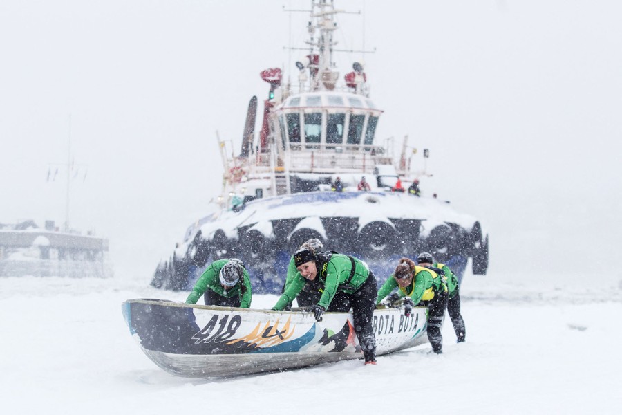A small crew of athletes push a canoe across a frozen river in front of a towboat.