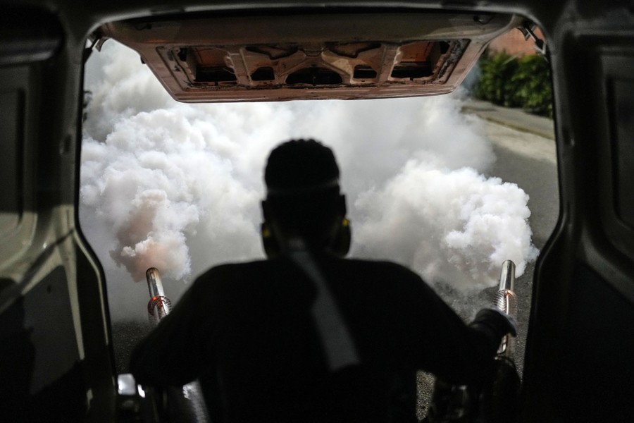 A person sits in a vehicle's open doorway, spraying clouds of pesticide over a road.