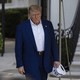 Donald Trump walking, looking displeased, carrying in one hand a hat that reads "USA"