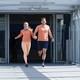 A still from the TV show The Amazing Race featuring a couple team in light orange athletic clothing running through an airport terminal
