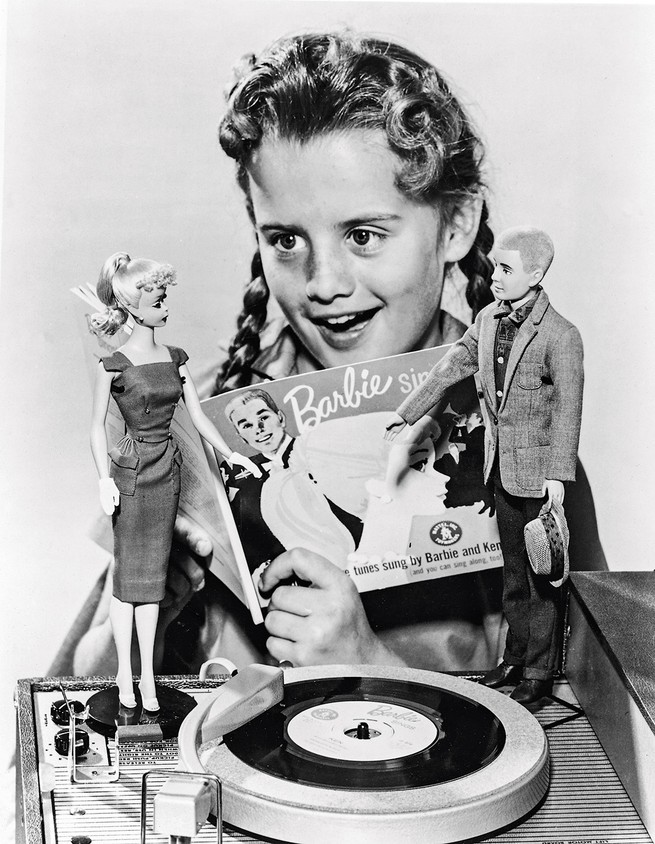 black and white photo of girl in braids holding "Barbie" booklet next to turntable with Barbie and Ken dolls
