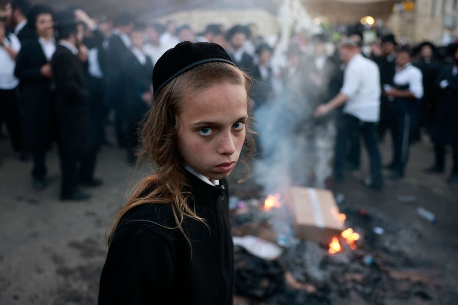 A young person looks at the camera as ultra-Orthodox Jews in the background stand around a fire.
