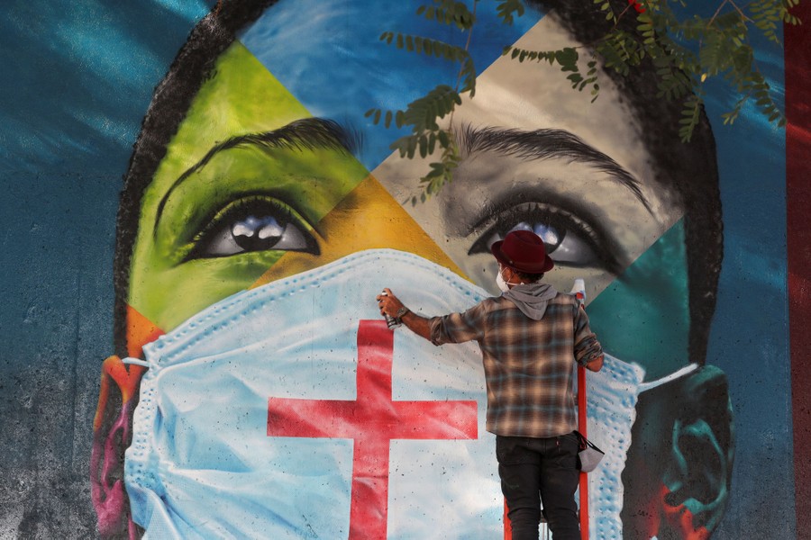 An artist stands in front of a large mural of the face of a young person wearing a mask.