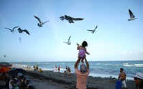 On a beach, a father holds his daughter up above his shoulders, and she looks up at the birds