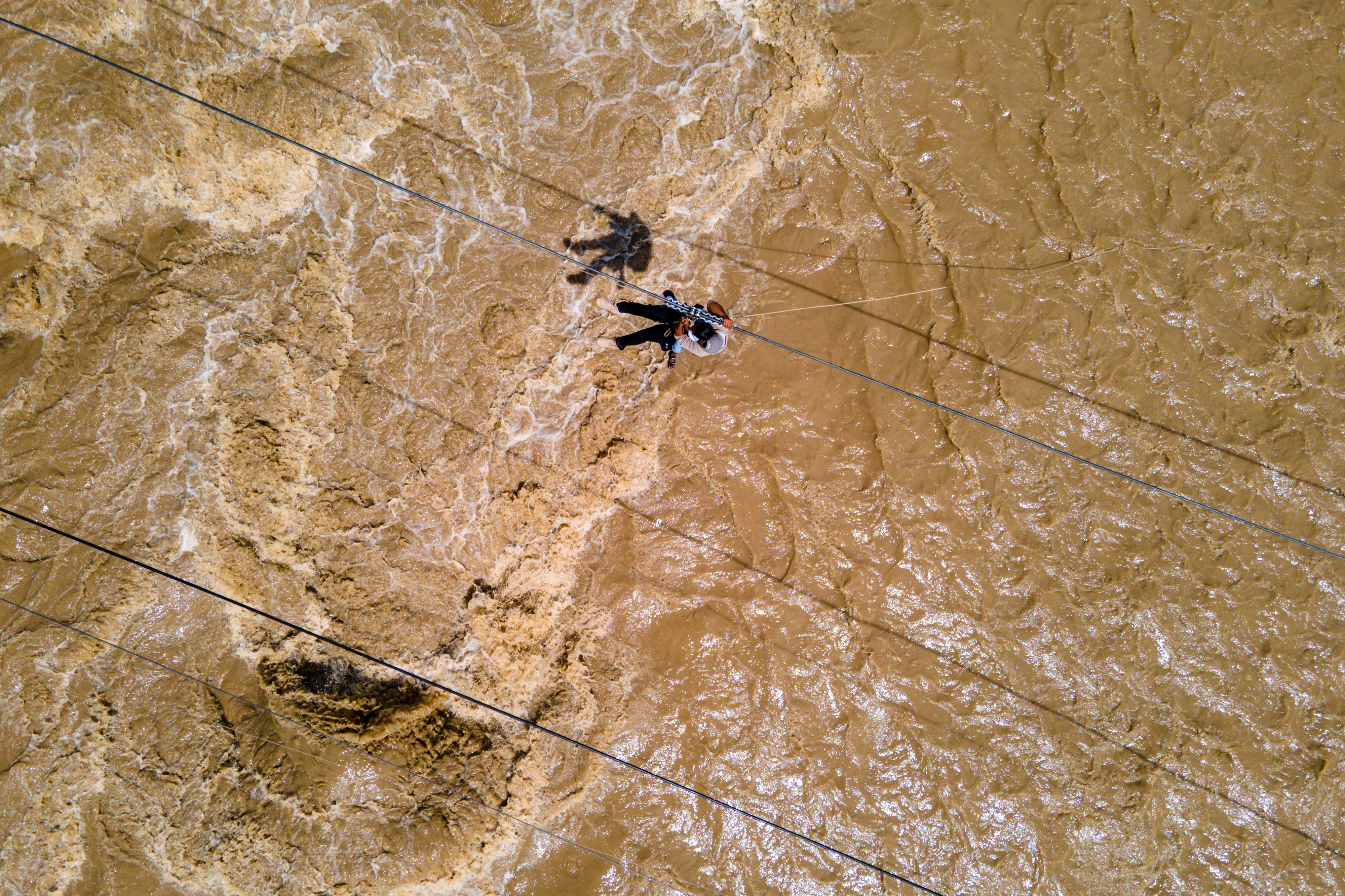 An aerial view of a person using a pulley system on a cable to cross over turbulent flood water.