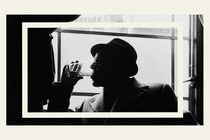 A black-and-white image of a man in side-profile wearing a hat and a coat, drinking from a clear glass