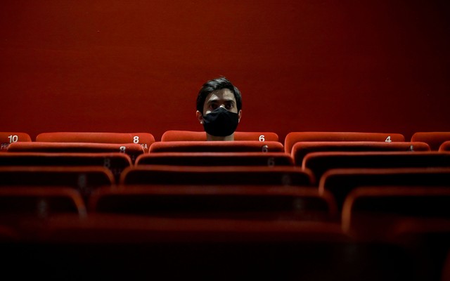 Person sitting alone in a red theater wearing a black mask