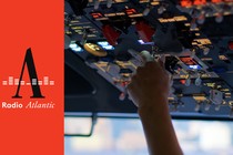 A pilot's hand extends up from the cockpit seat towards a ceiling full of switches. A pink manicured thumb nail is revealed and reaches towards a switch.