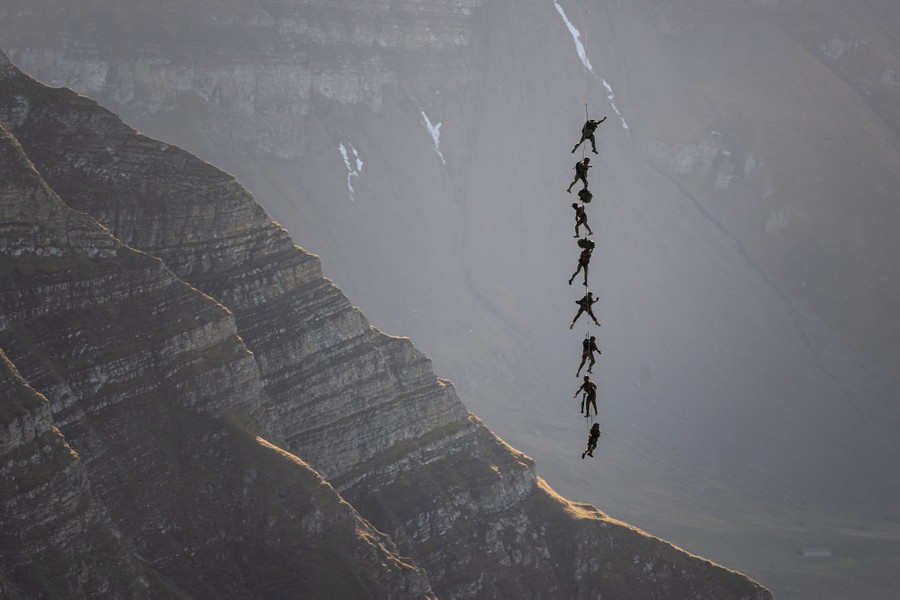 Eight soldiers dangle from a long cable, spaced out one above the other, flown through a mountain valley by a helicopter (not seen).