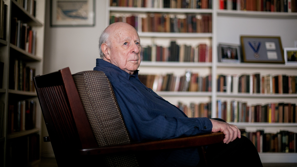 A photograph of an older Norman Podhoretz sitting in an armchair in a home library