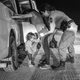 A black-and-white photo of a woman in a T-shirt and jeans leaning with both hands against a white van at night while a woman in uniform frisks her and a toddler stands underneath crying
