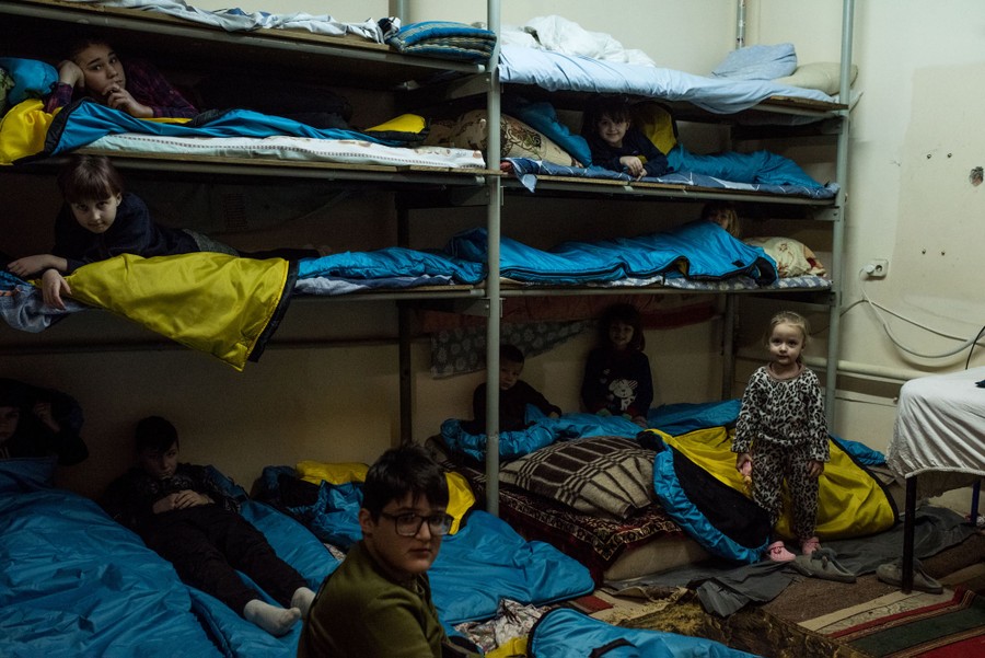 About nine children are seen, laying and sitting among blankets and bunk beds in a shelter.
