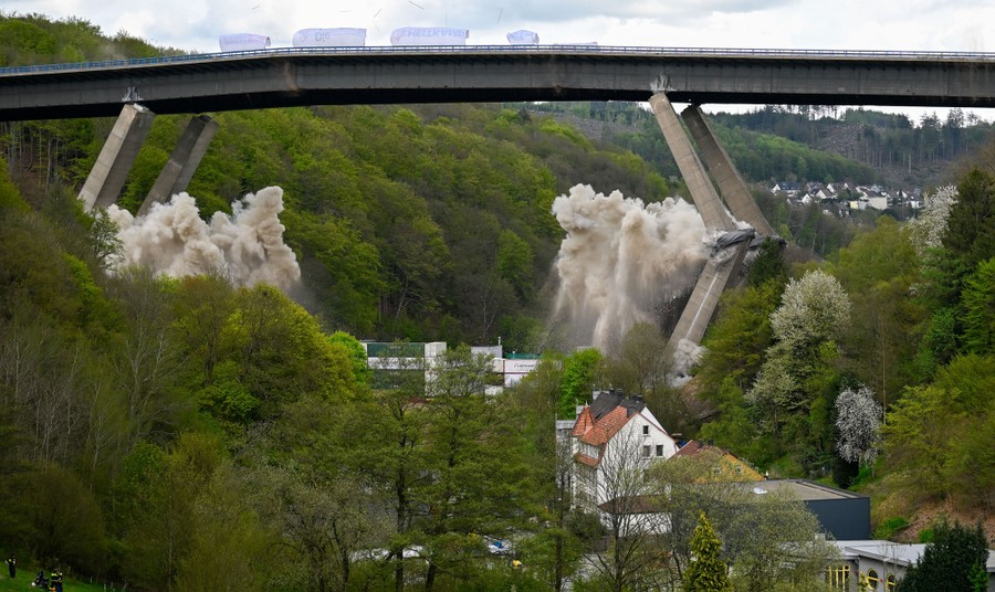 Explosives destroy supporting pillars beneath a highway bridge, which drops into a valley.