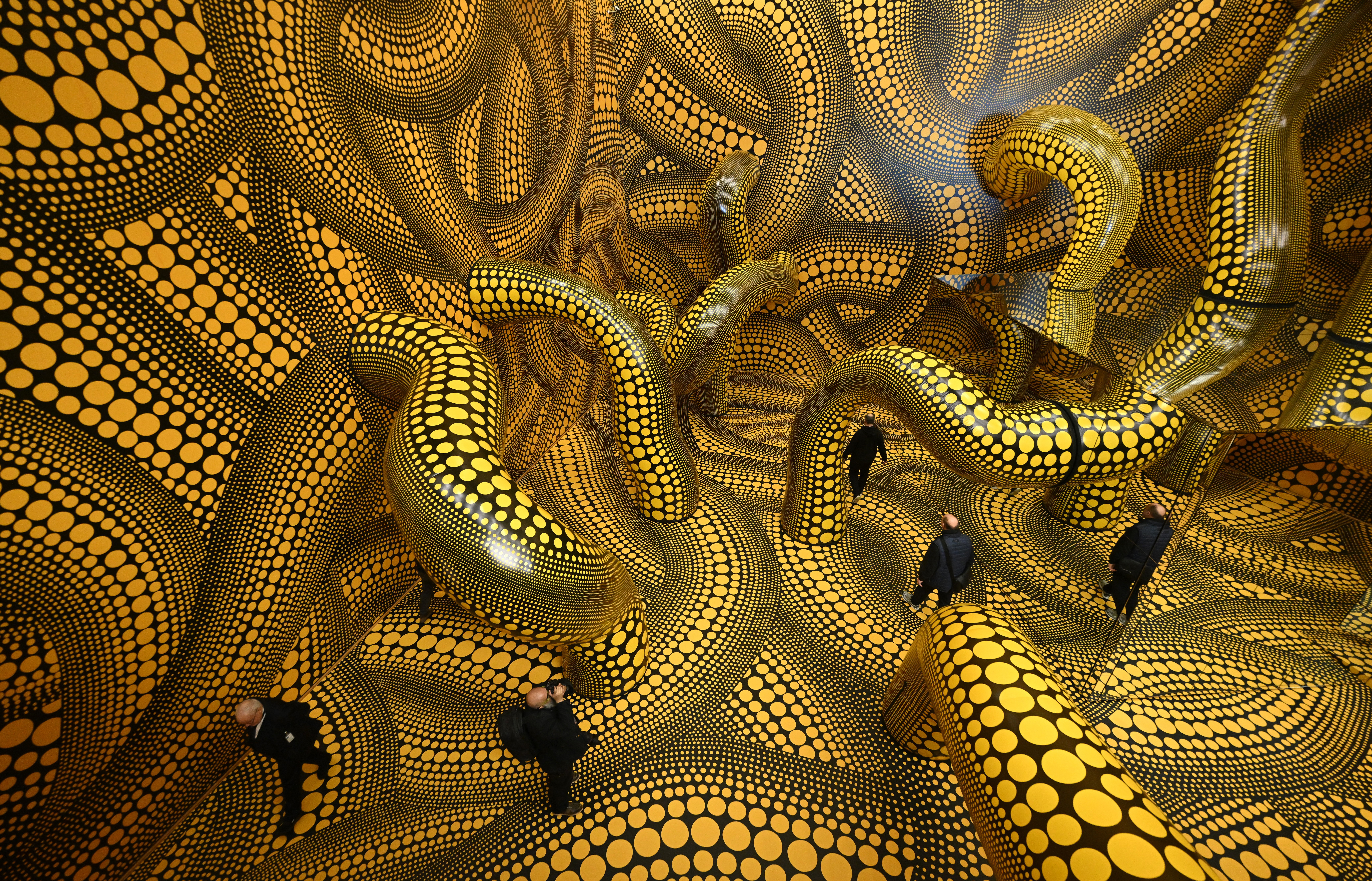 Visitors walk through a large room-sized art installation resembling dozens of twisting tubes decorated with yellow circles on black backgrounds.