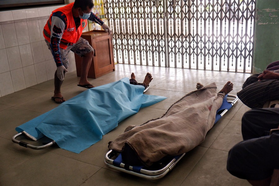 A person stands beside two bodies that are covered by blankets, on a tile floor.