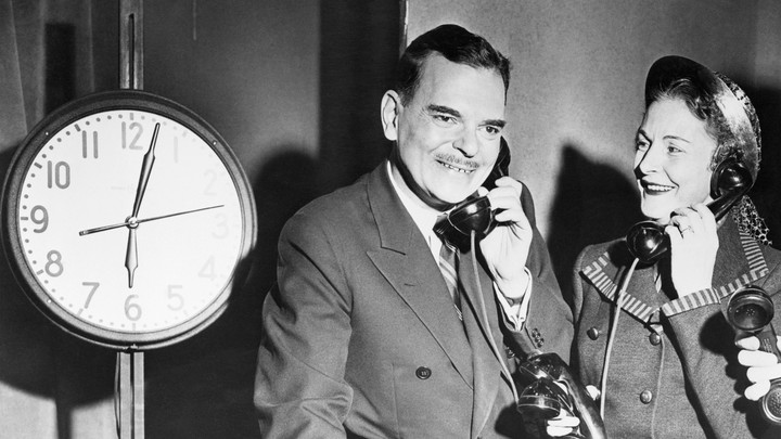 Two people talk on phones in front of a clock.