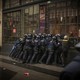 A group of riot police pushes together against a large door with protesters on the other side.