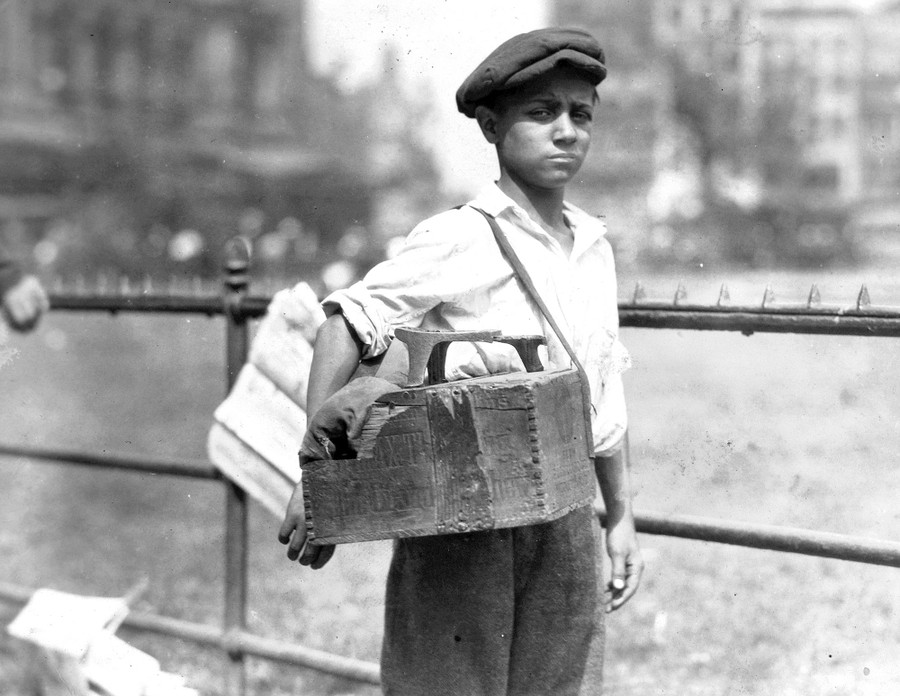 A young person stands in a park, with a shoe-shine box slung over their shoulder.