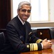 Surgeon General Vivek Murthy, in uniform, grins widely and clasps his hands on a shiny round table