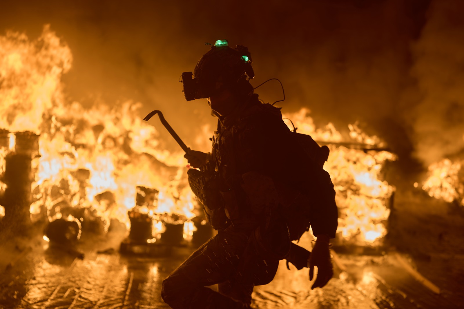 A firefighter carries a pry bar, walking past many burning objects after a missile attack.