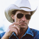 photo of Shepard in blue denim western shirt, cowboy hat, and sunglasses with hand raised to face