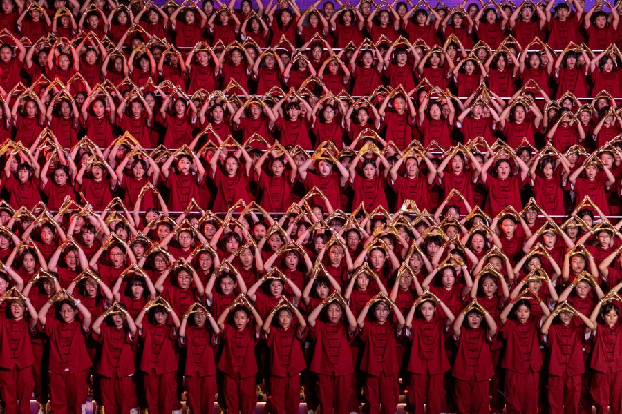 Dozens of students in red costumes stand side by side, holding their arms up, during a performance.