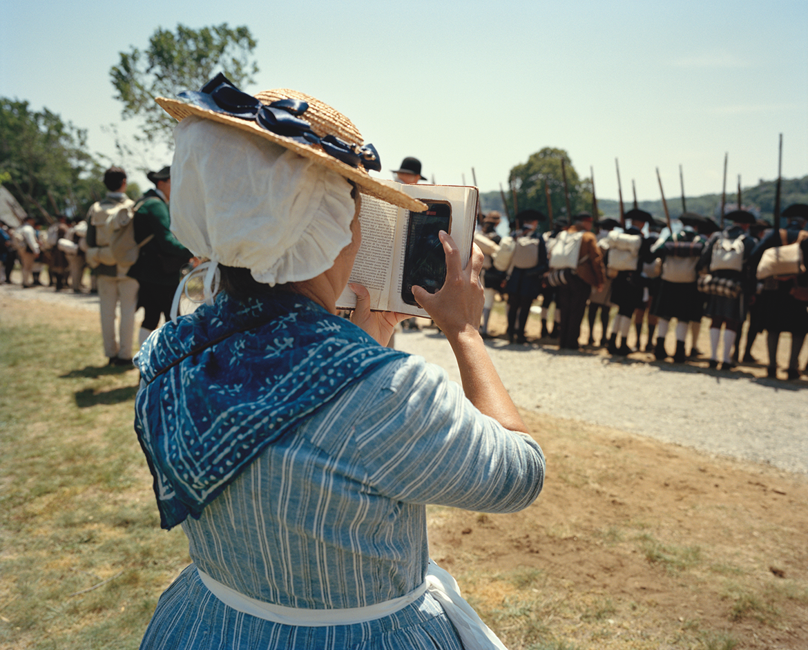 photo of female reenactor dressed in period blue dress, kerchief, and bonnet holding up a book that disguises the cell phone she's looking at behind it, with a row of soldiers with muskets facing away in the background