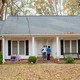 Color photograph of two people knocking on the front door a white and red-brick house with fallen leaves in the yard.