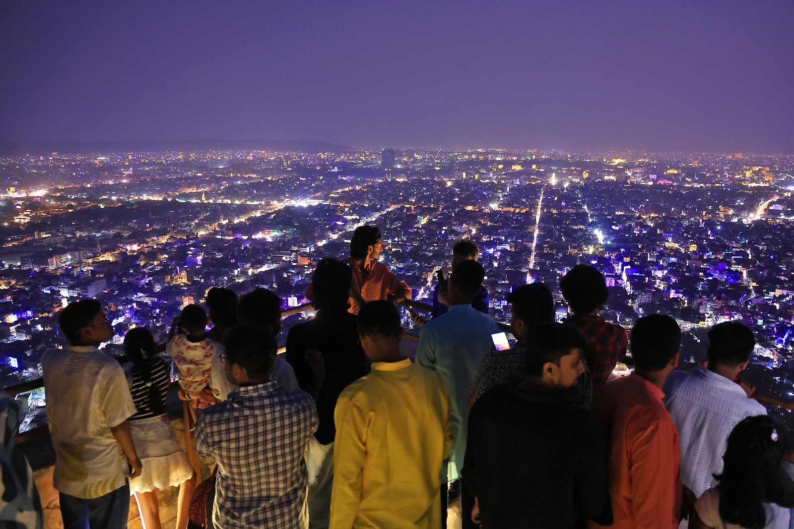 People stand in an observation area, overlooking a brightly illuminated city below.