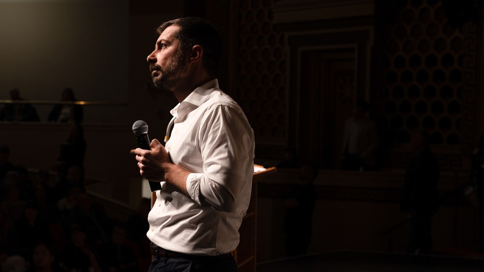 An image of Pete Buttigieg on a stage, holding a microphone, looking into a spotlight.