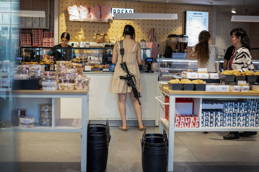A woman in civilian clothing carries a rifle on a shoulder strap while shopping in a bakery.