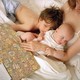 A toddler and baby lay in their mother's lap while she reads a book