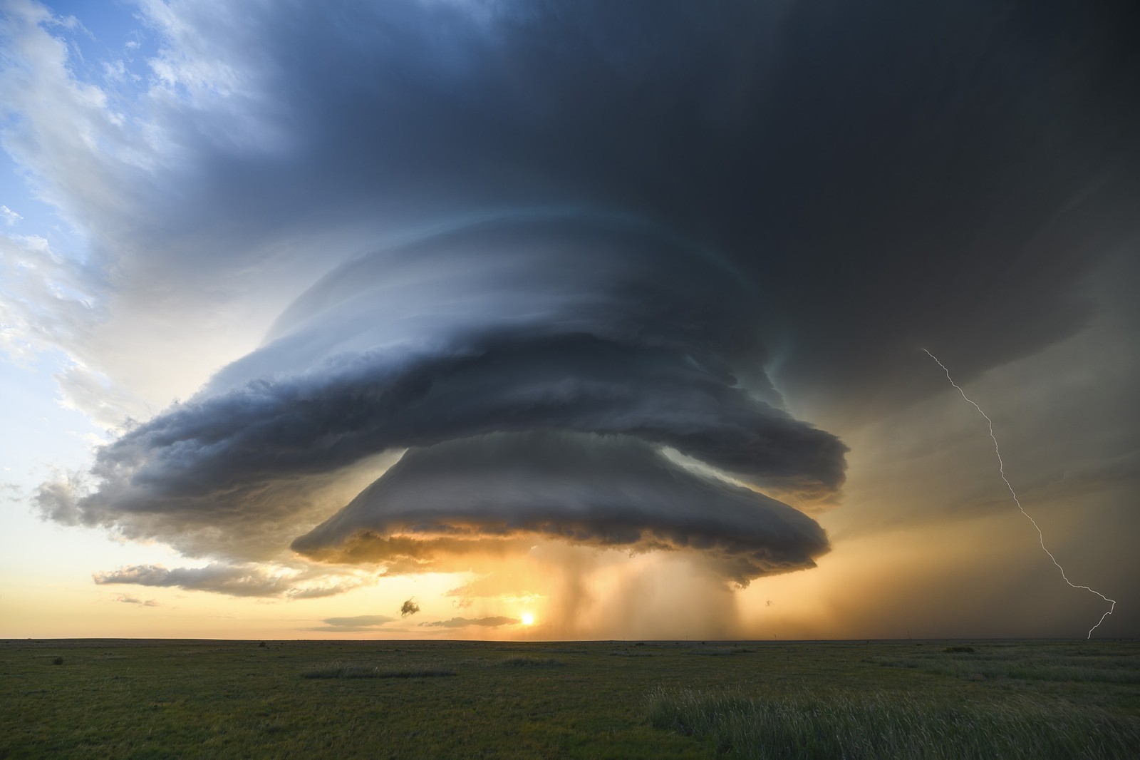 A massive storm cloud shaped like an upside-down funnel looms over farmland on a broad plain.