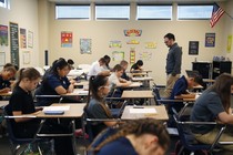 Students take a test in a classroom; a teacher stands at the front of the room. 