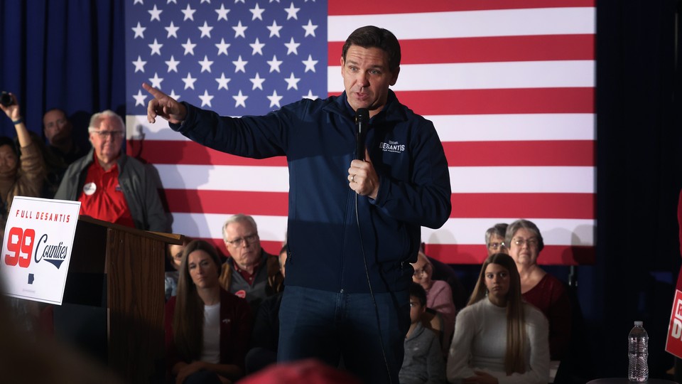 A photo of Ron DeSantis standing in front of a small, seated crowd and an American flag