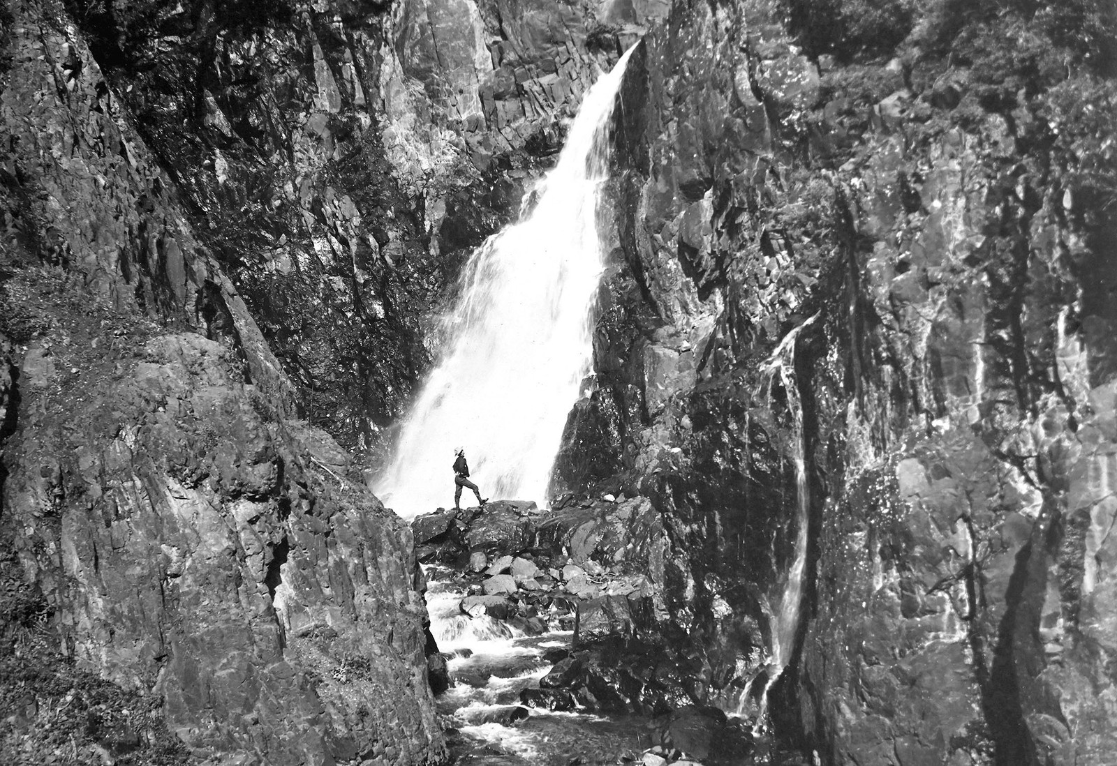 A person poses while standing beneath a tall waterfall.