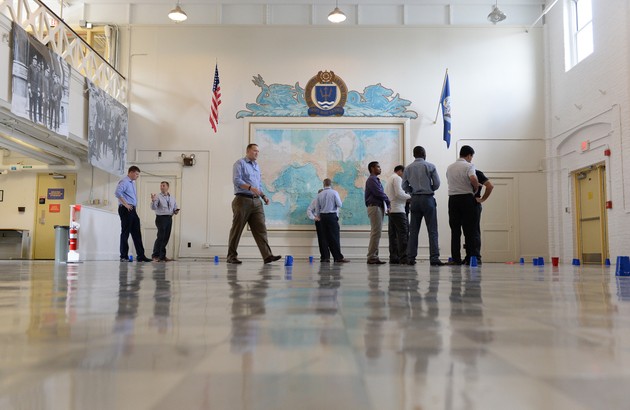 A group of men walks around a large room with a giant world map on the wall. Plastic cups rest upside down on the floor around the room.