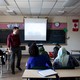 A teacher stands at the front of a classroom in Canada; students sit at desks, a few of whom are wearing hijabs, one of whom has dreadlocks. 