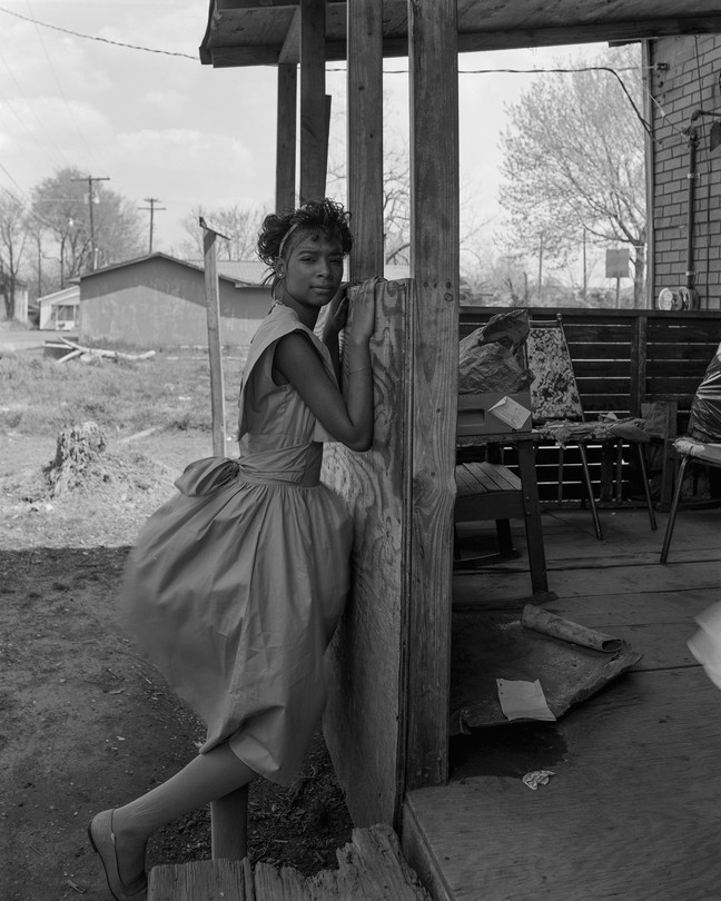 A young woman stands outside a wood house with her hands on the porch frame looking at the camera