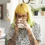 A young woman with dyed hair and tattoos sips a cup of coffee.
