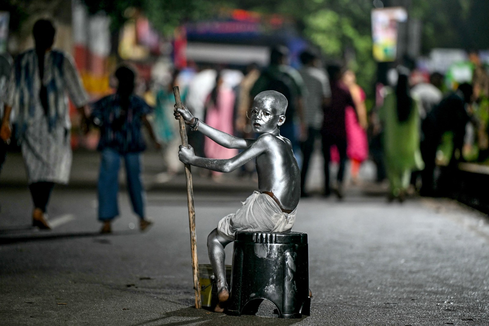 A boy, completely covered in silver paint, sits in a street while begging.