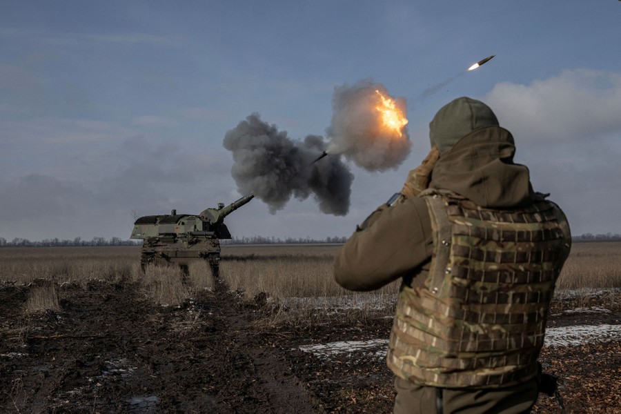 A soldier stands near a large piece of artillery as it fires a projectile.