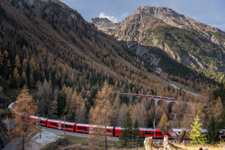 Një tren shumë i gjatë pasagjerësh udhëton në një rrugë të lakuar në një luginë të pjerrët malore.