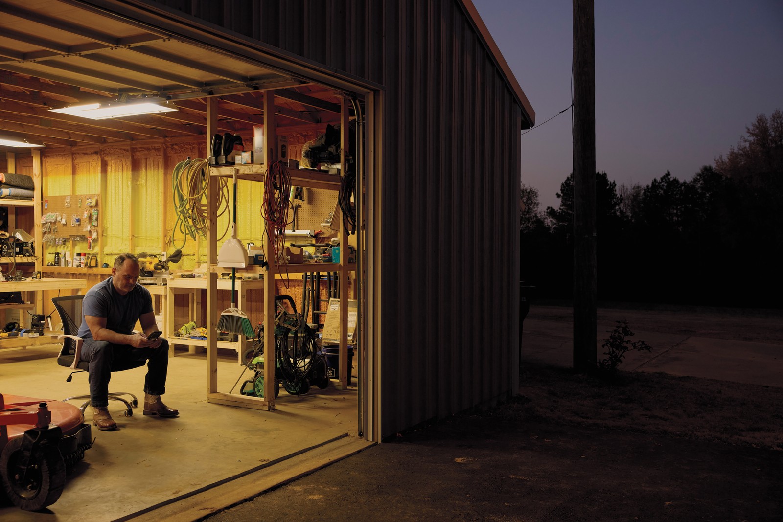 photo of man sitting in doorway of large well lit garage looking at phone at dusk