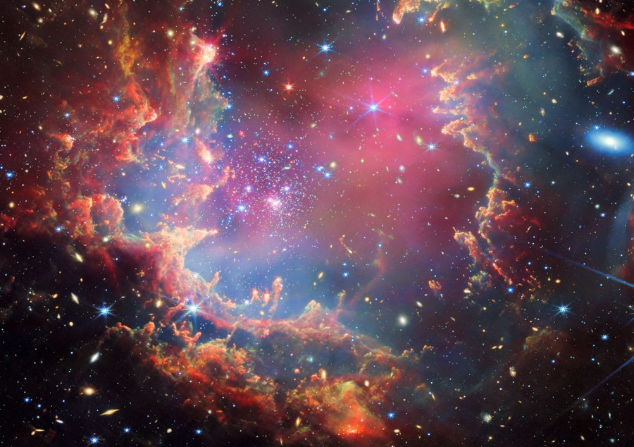 A star cluster is shown inside a large nebula of many-colored gas and dust. The material forms dark ridges and peaks of gas and dust surrounding the cluster, lit on the inner side, while layers of diffuse, translucent clouds blanket over them.
