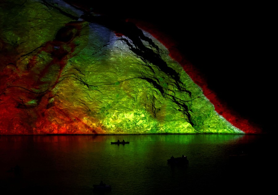 People paddle in small boats in front of light projections on a mountainside.