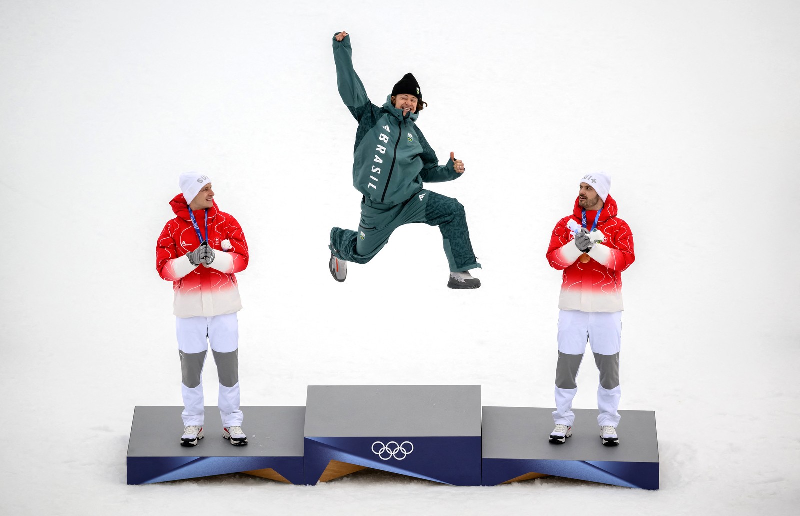 A skier jumps up, fist held high, smiling, as he climbs atop a podium to receive a gold medal, flanked by the bronze and silver winners.