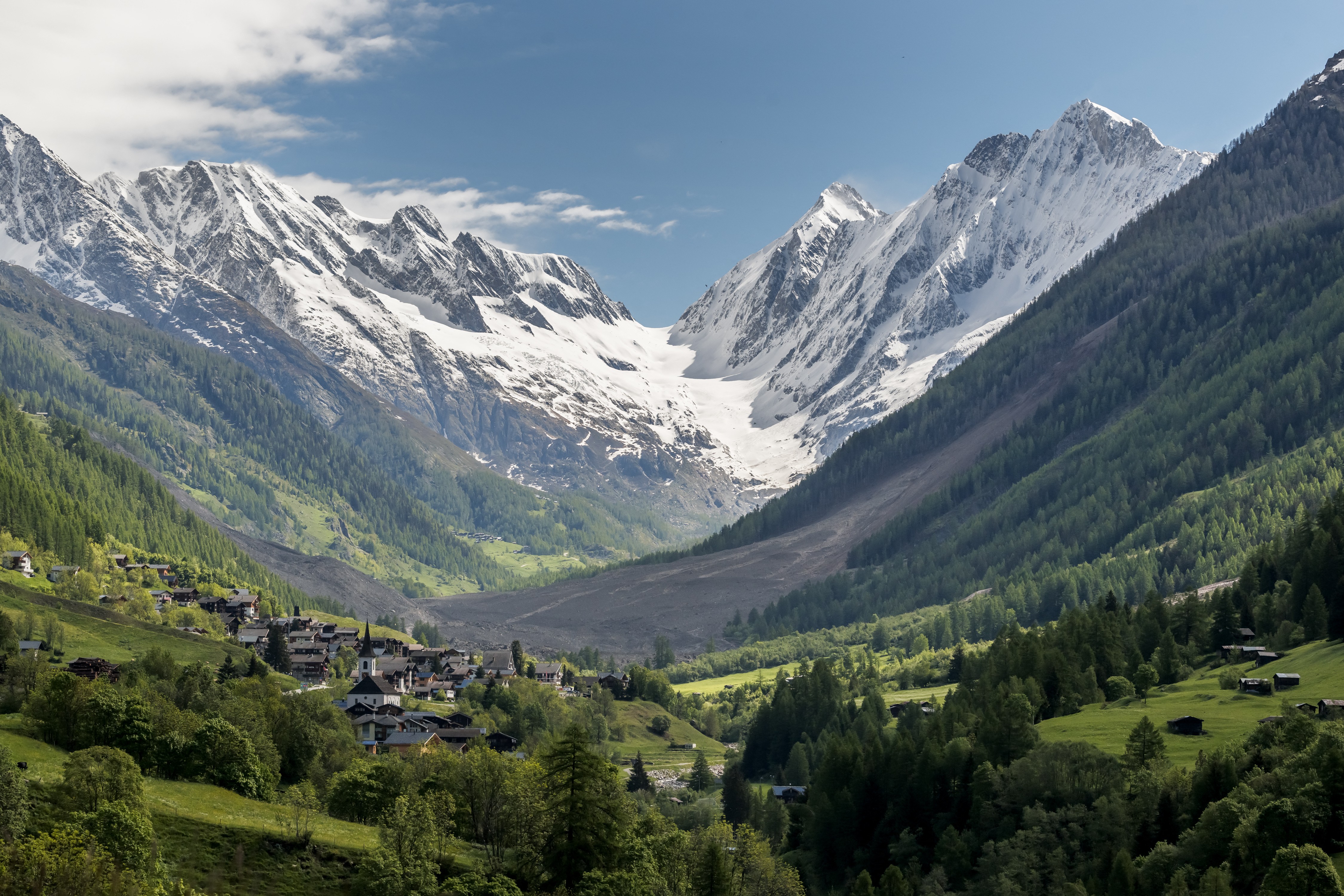 A broad view of a narrow mountain valley with a landslide at center, and some village buildings in the foreground
