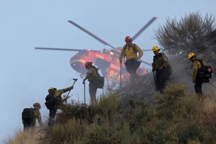 A firefighting helicopter flies above a hill where firefighters are at work.