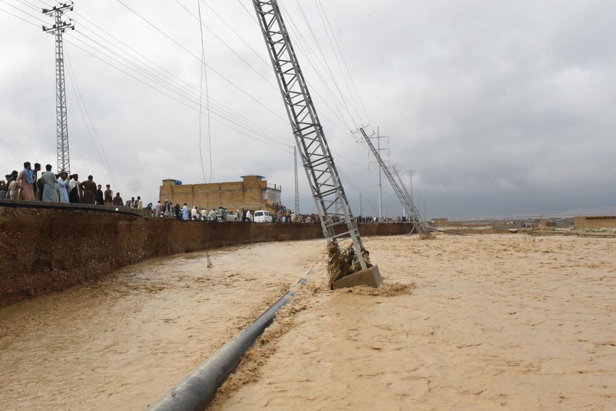 People stand at the edge of a heavily eroded roadway, watching floodwater pass by.